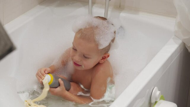 toddler is playing in the bath and holding a grey bottle of soap. They are surrounded by bubbles and appear to be having fun.