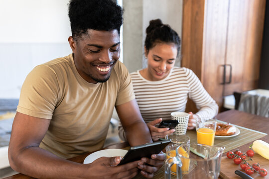 Happy couple using a tablet and phone during breakfast, enjoying a relaxed morning together in a bright and cozy kitchen setting - Powered by Adobe