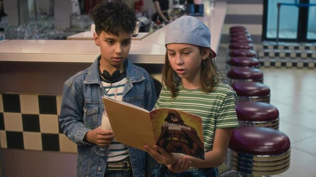 Medium shot of two diverse teenage kids standing in retro cafe, Indian boy in denim jacket holding milkshake, Caucasian girl in baseball cap with comic book, looking at pictures and discussing plot