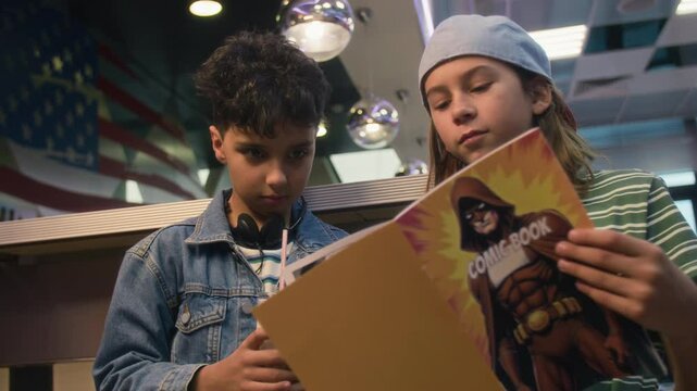 Medium shot of two multiethnic young boys leafing through comic or anime magazine in American diner, Caucasian kid talking about story and pointing at favourite heroes, Indian boy drinking milkshake