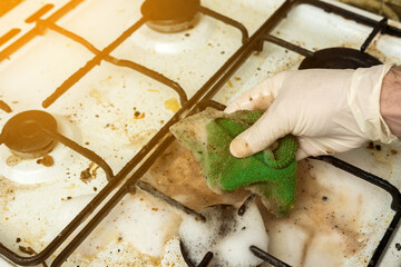 Foamy soap cleaning a dirty gas stove burner, with a hand in gloves using a green cloth. A representation of household chores and kitchen maintenance