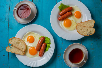 Breakfast for two fried eggs with sausages crispy white bread and tea in cups on a blue