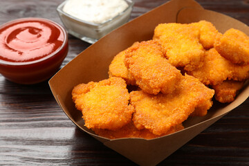 Tasty chicken nuggets in paper container and sauces on wooden table, closeup