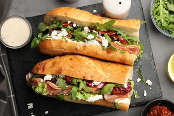 Halves of baguette sandwich with prosciutto and sun dried tomatoes and ingredients on grey table, flat lay