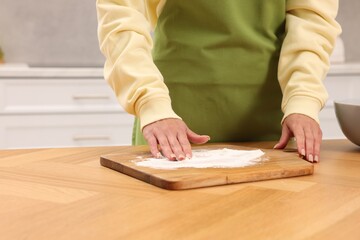Woman sprinkling flour onto wooden board before rolling dough at table indoors, closeup