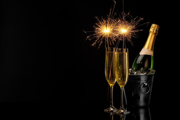 Glasses of sparkling wine with sparklers and bottle in cooler bucket on black background. Space for text