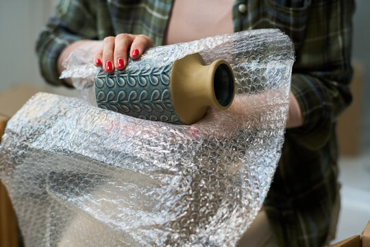 Hand of unrecognizable young woman wrapping vase into bubble wrap before packing it into box while preparing for relocation