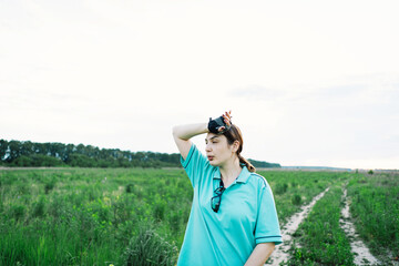 Woman in a blue shirt standing in a field with her arms crossed. She is wearing cycling gloves and has sunglasses hanging from her shirt. The woman appears to be resting after a bike ride.