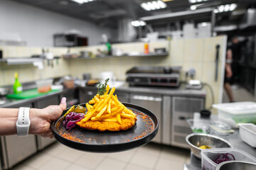 A close-up of a hand presenting a plate featuring golden fries and a breaded chicken fillet, set against a busy restaurant kitchen background. Bright colors and appetizing textures.