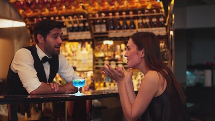 Sitting in smiling beautiful woman guest lively taking with smart bartender while drinking mixed vodka in blue hawaii cocktail and whiskey at luxurious counter bar on Friday meeting at club. Vinosity.