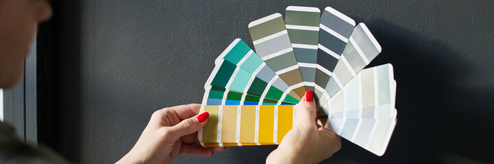 Header of hands of young woman holding color palette against grey painted wall of living room...