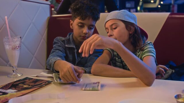 Medium shot of two bored young diverse 1980s teenage boys sitting together at table in cafe at entertainment arcade, counting game coupons and pocket money dollars, discussing weekend plans