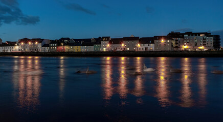 Late evening view of The Long Walk in Galway, with swans gliding in motion blur on the river.