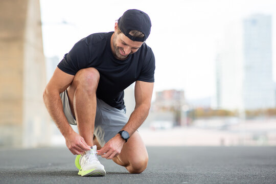 Man athlete in sportswear crouching and tying shoelaces outdoors