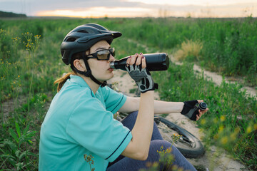 A woman in a bike helmet and sunglasses takes a drink from a reusable water bottle while out on a bike ride. She is in a field with green grass and a cloudy sky above.