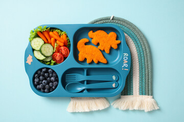 Fototapeta premium Kids meal in a blue divided plate with dinosaur-shaped chicken nuggets, fresh vegetables, and blueberries, placed on a blue and white woven mat. Fun and healthy food presentation for children