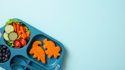 Children's meal with dinosaur-shaped chicken nuggets, fresh vegetables, and blueberries in a blue divided plate on a light blue background