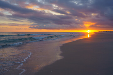 sun sets in a dramatic explosion of colors over a peaceful beach, with the waves gently breaking on the shore and figures in the distance