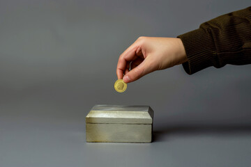 A hand places a coin into a metal box against a gray background. Concept of saving or donation. For financial advice.