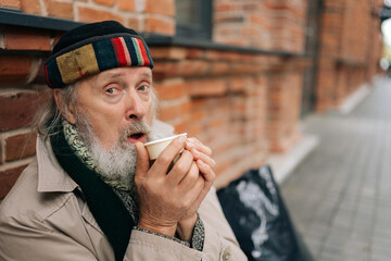 Closeup of elderly man sitting outside on city street, sipping warm drink from paper cup, evoking...