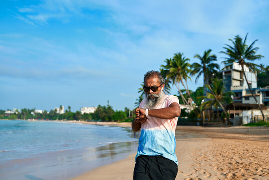 Senior ethnic man on beach uses smartwatch and wireless earbuds. Bearded old grandpa embraces modern tech innovations. Active lifestyle with wearable gadgets. Ocean backdrop creates calm vibe.