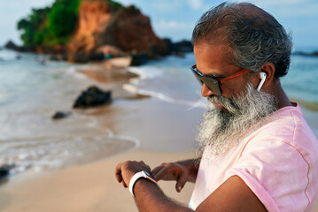 Senior ethnic man uses smart watch, wireless earbuds on tropical beach. Bearded elderly enjoys modern tech for fitness, wellbeing. Healthy lifestyle enhances training. Mature grandpa embraces gadgets.