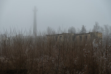 Abandoned and weed-covered industrial area with a building and a tall chimney in winter