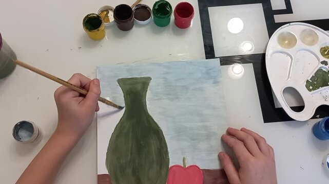 a left-handed girl draws a gouache vase on a white sheet of paper on a glass table, top view, her hands are visible