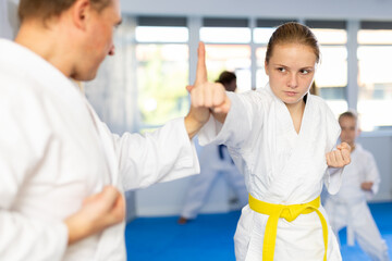 Obraz premium Young girl in kimono and colored belt practicing karate punch block during group martial arts lesson in gym, accompanied by trainer