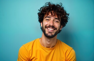 Obraz premium Cheerful young man poses for photo. Indoor portrait against blue background. Happy expression. Attractive dark-haired guy with curly hair, beard. Posing for camera with toothy smile. Wearing yellow