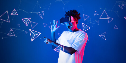 Smart boy standing at blue background while floating geometric shape. African man playing game and wearing VR goggle and casual shirt. Augmented reality technology innovation concept. Ingenuity.
