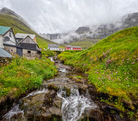Obraz premium Bucolic scene of a Scandinavian village in the majestic mountains. A stream of water flows down from the mountains. Low clouds and fog hang over the scene. Kunoy, Faroe Islands, Kingdom of Denmark.
