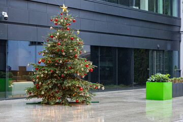 Beautifully decorated Christmas tree adorned with red and gold ornaments, glowing lights and a golden star on top, standing outdoors near a modern building during light drizzling rain on a winter day