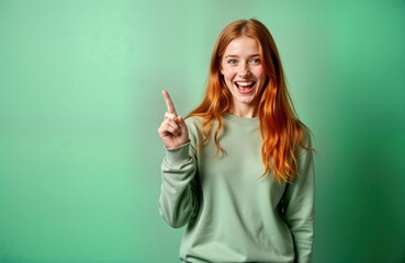 Fototapeta premium Excited ginger teen girl points upwards with happy expression. Young redhead female model stands isolated studio green background. Casual clothing. Wow emotion. Positive attitude. Attractive teen.
