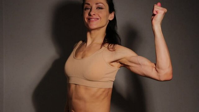 Healthy smiling woman posing and showing strong strength biceps muscle, attractive body. Slim caucasian female model posing in beige bra in studio low key light on wall background. Sporty lifestyle