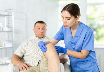 Middle-aged male patient lying on clinical chair having his knee examined by young doctor