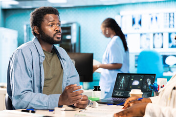 Black male patient bringing x ray test results to medic for scan interpretation, reviewing the reports for diagnostics. Medic expert evaluating the radiography film in cabinet.