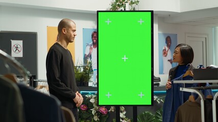 Asian store assistant next to green screen presenting the new clothes collection to client, ensuring a great customer service and helping young man with fashion advice. Retail business.