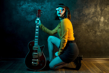Stylish young hipster woman with curly hair with red guitar in neon lights. Rock musician posing...