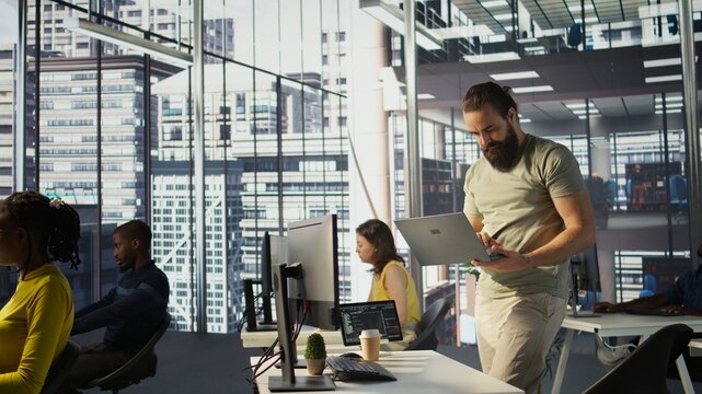 Admin at office desk utilizing cloud computing resources on laptop to train large scale machine learning models. Man monitoring company systems using notebook while standing in workspace, camera B