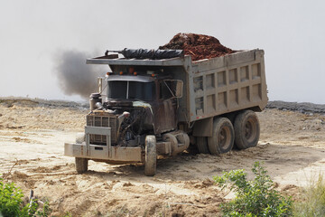 A dump truck loaded with soil is navigating through a construction site, surrounded by raw earth, as smoke billows from its exhaust.