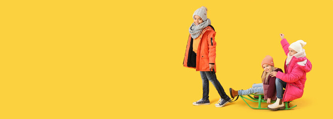 Cute children in winter clothes with sledge on yellow background