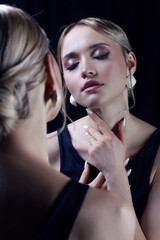 Portrait of young blonde woman near mirror with eyes closed. Female in black dress with straps and earring.