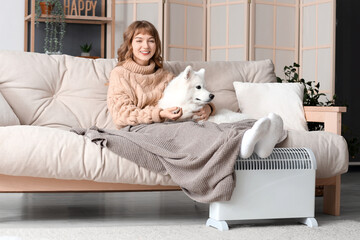 Young woman with blanket and Samoyed dog on sofa warming near radiator at home