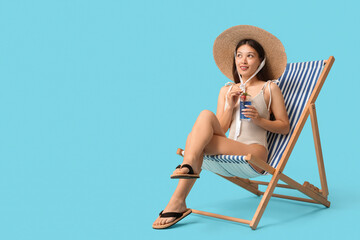 Beautiful young Asian woman in swimsuit with glass of cocktail sitting in deck chair on blue background