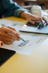 Close-up of a person working on a laptop, analyzing financial graphs and data with a pen in an office environment.