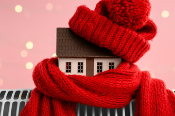 Model of house with warm scarf and hat on electric heater against blurred lights background. Concept of heating season