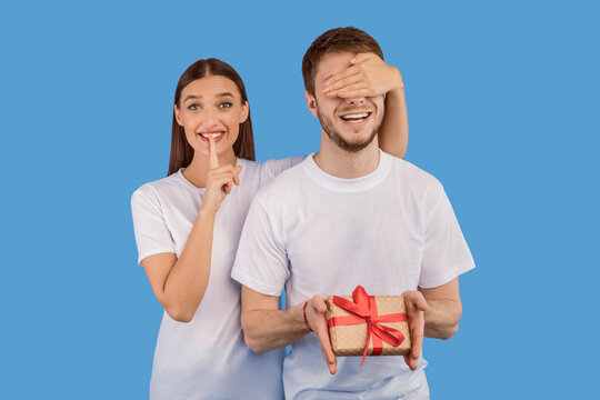 It's A Secret. Girl covering her boyfriend eyes, making surprise and silence gesture, isolated over pastel purple studio wall