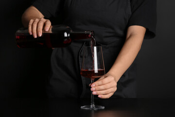 Female sommelier pouring red wine from bottle into glass on dark background