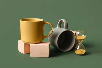 Composition with mugs, plaster podiums and hourglass on green background